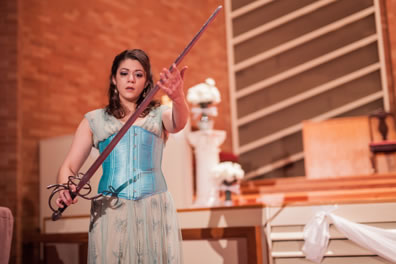 Aspatia in a filly blue patterned cream dress and pastel blue corset strokes a long sword she is holding in her hands. In the background is the sancuary of the church