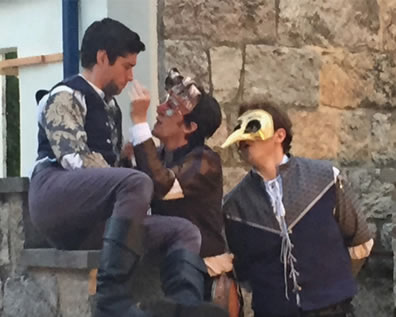 Romeo sits on a step, in blue waistcoat, bue floral patterned shirt, purple pants and black boats as Mercutio, mask perched on his head and in brown checked shirt, tweaks his nose. Benvolio, wearing a long-nose mask and blue and gray jerkin, stands behind Mercutio, with the stone walls behind them.