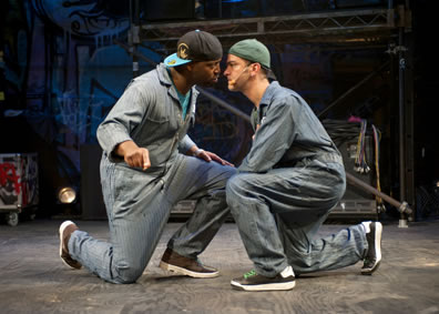 Squatting on a knee, Othello and Iago in coveralls, baseball hats worn backward, and using microphone headsets, get face to face,