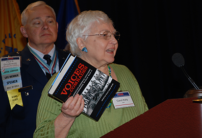 Carol_Book_ROA Carol Kelly in olive green dress holds "Voices of our Comrades" in her hands as she speaks at the podium as retired Air Force colonel Paul Groskreutz in his service uniform watches behind her