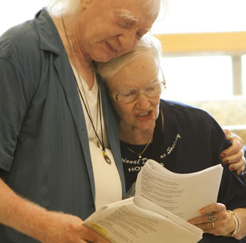 Condos with blue shirt unbuttoned and a white t-shirt underneath, necklaces hanging down around his neck and long white hair hugs Fairchild to his breast with his left arm around her shoulder, she wearing wireframe glasses and a black t-shirt as she reads a script; both hold scripts in their other hand.