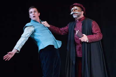 Shylock in a red quilt-like tunik, black academic robe and red hat and wearing a brown mask with large white mustache and shaggy eyebrows holds a small account book in left hand and grabs the aqua blue tunic of Bassanio, who leans backward with arms spread.
