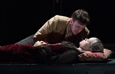 Romeo in gold jacket and jeans leans over Juliet asleep on a bier, head on embroidered pillow, and she's wearing a velvet red dress with black sleeves