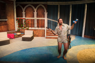 Benedick in tan beach shirt and swim trunks with sunglasses in one hand and blue bottle of lotion in the other with a folded up beachchair hanging over his shoulder.