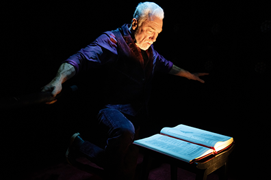 Patrick Page in All the Devils Are Here 6 Patrick Page in dark purple shirt and black vest and pants, his face glowing in a spotlight, has his arms outstretched as he leans over an open book on the table.