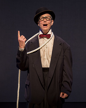 Bill Irwin wearing a black dinner jacket, black baggy pants, white shirt, red bow tie, large black frame glasses, and a black bowler hat has a rope noose around his neck with the rope going down toward the floor. His right hand's index finger is pointing up.