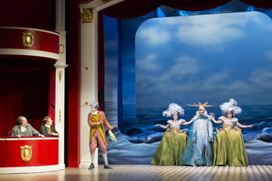 Dangle and Sneer sit in red theater box seats to the left, Puff stands near them talking to them, and to his left in the center of the stage are two women dressed as shorelie (green wide hoops skirts with sillhouettes of towns on the hips and clouds for hats) and a man dressed as an ocean with sun rays for a hat. Behind them is the English channel, a mechanism emulating waves.